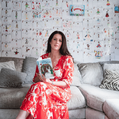 clare anderson sitting on grey sofa in red dress holding the lost wish book