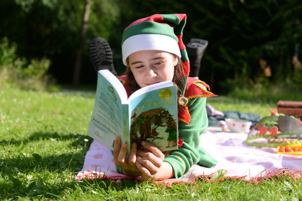 young girl wearing elf hat reading a book whilst laying on the grass